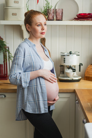 Pregnancy motherhood people expectation future. Pregnant woman with big belly smiling in kitchen at home. Girl hugging her tummy enjoying pregnancy. Maternity tenderness parenthood new life conceptの写真素材
