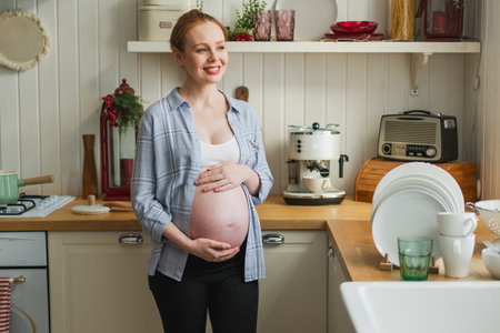 Pregnancy motherhood people expectation future. Pregnant woman with big belly smiling in kitchen at home. Girl hugging her tummy enjoying pregnancy. Maternity tenderness parenthood new life conceptの写真素材