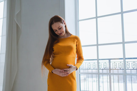 Pregnancy motherhood people expectation future. Pregnant woman with big belly standing near window at home. Girl hugging her tummy enjoying pregnancy. Maternity tenderness parenthood new life conceptの写真素材