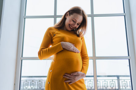 Pregnancy motherhood people expectation future. Pregnant woman with big belly standing near window at home. Girl hugging her tummy enjoying pregnancy. Maternity tenderness parenthood new life conceptの写真素材