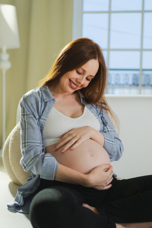 Pregnancy motherhood people expectation future. Pregnant woman touching big belly sitting on couch at home. Girl hugging her tummy enjoying pregnancy. Maternity tenderness parenthood new life conceptの写真素材