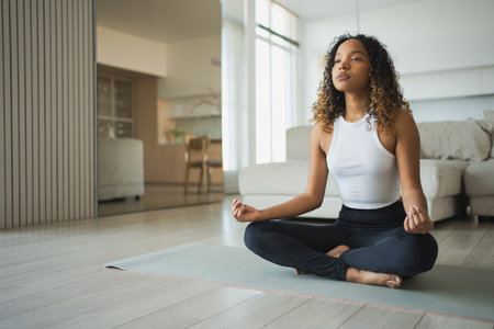Yoga mindfulness meditation. Young healthy African girl practicing yoga at home. Woman sitting in lotus pose on yoga mat meditating relaxing indoor. Girl doing breathing practice. Yoga at homeの写真素材