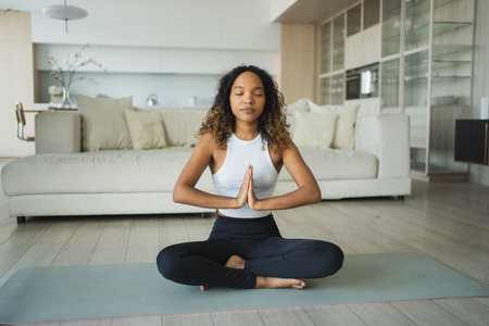 Yoga mindfulness meditation. Young healthy African girl practicing yoga at home. Woman sitting in lotus pose on yoga mat meditating relaxing indoor. Girl doing breathing practice. Yoga at homeの写真素材