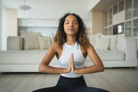 Yoga mindfulness meditation. Young healthy African girl practicing yoga at home. Woman sitting in lotus pose on yoga mat meditating relaxing indoor. Girl doing breathing practice. Yoga at homeの写真素材