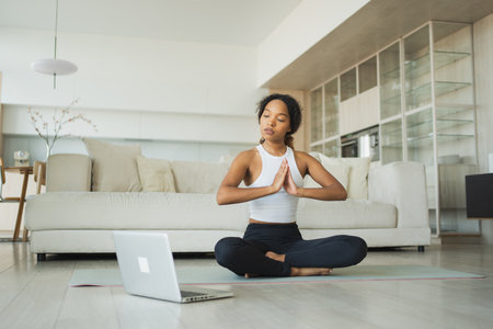 Yoga mindfulness meditation online. African woman practicing yoga with online lessons in laptop at home. Woman sitting in lotus pose meditating relaxing. Girl doing breathing practice. Yoga at homeの写真素材