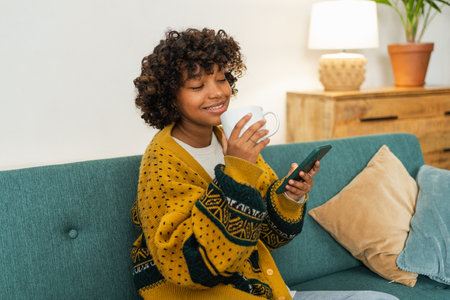 African girl drinking coffee holding smartphone sitting on couch at home Woman with cell phone surfing internet using social media apps. Shopping online Internet news cellphone addictionの写真素材