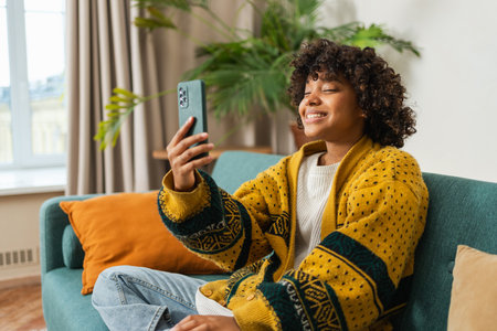 African American girl holding smartphone having video chat. Female blogger chatting with best friends in social network. Young woman having virtual meeting online chat video call at homeの写真素材