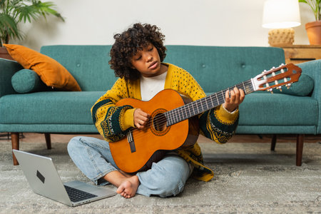 African American woman learning guitar watching tutorial on laptop at home. Black girl playing guitar singing song learning online music lessons. Artistic woman playing acoustic guitar learning chordsの写真素材