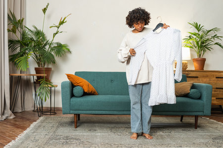Shopping fashion style. African woman choosing dress at home. Young woman trying to choose dress to wear holding hanger with clothes. Curly dark skinned girl smiling holding hanger with white dressの写真素材