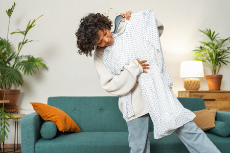 Shopping fashion style. African woman choosing dress at home. Young woman trying to choose dress to wear holding hanger with clothes. Curly dark skinned girl smiling holding hanger with white dressの写真素材