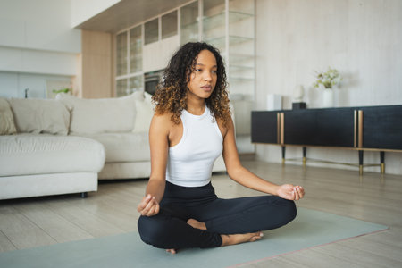 Yoga mindfulness meditation. Young healthy African girl practicing yoga at home. Woman sitting in lotus pose on yoga mat meditating relaxing indoor. Girl doing breathing practice. Yoga at homeの写真素材
