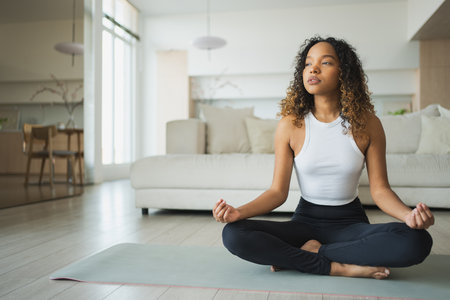 Yoga mindfulness meditation. Young healthy African girl practicing yoga at home. Woman sitting in lotus pose on yoga mat meditating relaxing indoor. Girl doing breathing practice. Yoga at homeの写真素材