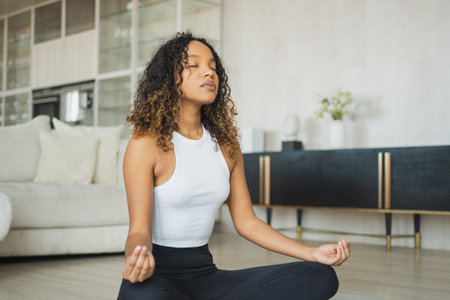 Yoga mindfulness meditation. Young healthy African girl practicing yoga at home. Woman sitting in lotus pose on yoga mat meditating relaxing indoor. Girl doing breathing practice. Yoga at homeの写真素材