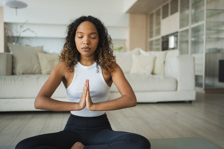 Yoga mindfulness meditation. Young healthy African girl practicing yoga at home. Woman sitting in lotus pose on yoga mat meditating relaxing indoor. Girl doing breathing practice. Yoga at homeの写真素材