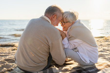 Senior mature couple sitting on beach holding hands enjoying outdoor recreation. Old husband wife touching hands with tenderness love. Grandmother grandfather together. Family moment of loveの写真素材