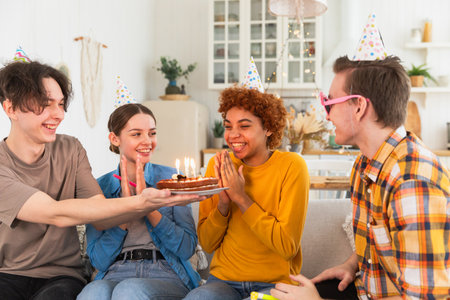 Make a wish. Woman wearing party cap blowing out burning candles on birthday cake. Happy Birthday party. Group of friends wishes girl happy birthday. People celebrating birthday with party at homeの写真素材