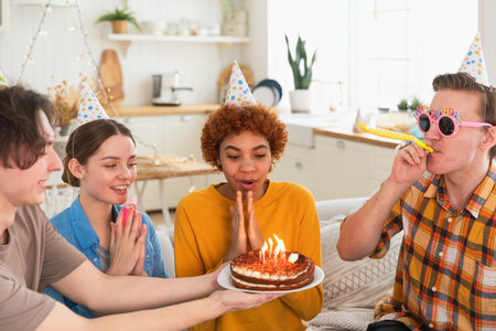 Make a wish. Woman wearing party cap blowing out burning candles on birthday cake. Happy Birthday party. Group of friends wishes girl happy birthday. People celebrating birthday with party at homeの写真素材