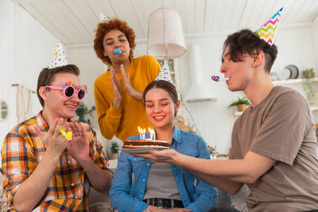 Make a wish. Woman wearing party cap blowing out burning candles on birthday cake. Happy Birthday party. Group of friends wishes girl happy birthday. People celebrating birthday with party at homeの写真素材