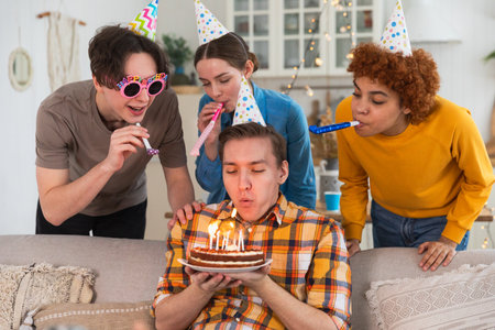 Make a wish. Man wearing party cap blowing out burning candles on birthday cake. Happy Birthday party. Group of friends wishes guy happy birthday. People celebrating birthday with party at homeの写真素材