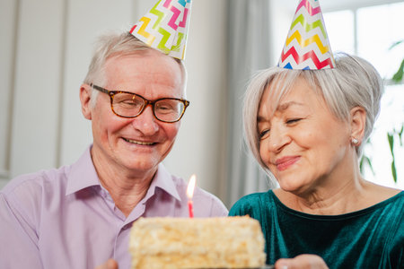 Make a wish. Family senior couple in party hat cap celebrating birthday anniversary together at home. Old woman blowing out burning candles on birthday cake. Old man wishes wife happy birthdayの写真素材
