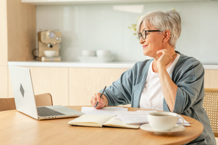 Middle aged senior woman using laptop computer writing notes at home. Focused mature old businesswoman work in home office making written records doing online work. Mature boss professional workerの写真素材