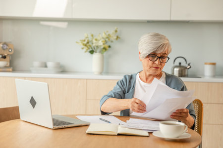 Middle aged senior woman sit with laptop and paper document, Pensive older mature lady reading paper bill pay online at home managing bank finances calculating taxes planning loan debt pension paymentの写真素材