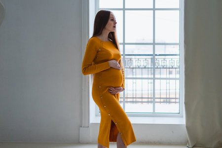 Pregnancy motherhood people expectation future. Pregnant woman with big belly standing near window at home. Girl hugging her tummy enjoying pregnancy. Maternity tenderness parenthood new life conceptの写真素材