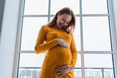 Pregnancy motherhood people expectation future. Pregnant woman with big belly standing near window at home. Girl hugging her tummy enjoying pregnancy. Maternity tenderness parenthood new life conceptの写真素材