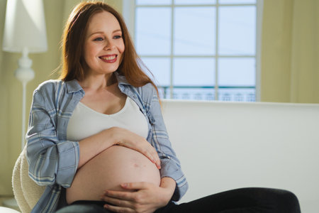 Pregnancy motherhood people expectation future. Pregnant woman touching big belly sitting on couch at home. Girl hugging her tummy enjoying pregnancy. Maternity tenderness parenthood new life conceptの写真素材