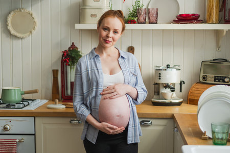 Pregnancy motherhood people expectation future. Pregnant woman with big belly smiling in kitchen at home. Girl hugging her tummy enjoying pregnancy. Maternity tenderness parenthood new life conceptの写真素材