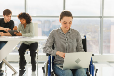 Accessible environment. Woman in chair for people with disability working with laptop in modern office. Diverse multiracial group of people colleagues working together. Young professional teamの写真素材