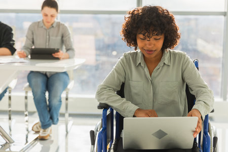 Accessible environment. African woman in chair for people with disability working with laptop in modern office. Diverse multiracial group of people colleagues working together. Young professional teamの写真素材