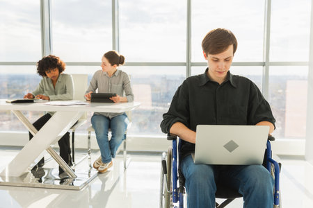 Accessible environment. Young man in chair for people with disability working with laptop in modern office. Diverse multiracial group of people colleagues working together. Young professional teamの写真素材