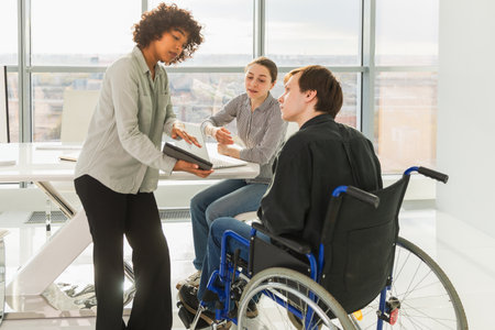 Teamwork brainstorm. Diverse multiracial colleagues man in chair for people with disability working together. Group of people sitting in creative office discussing business. Young professional teamの写真素材