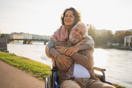 Old man in wheelchair walking with caregiver senior woman on road in park. Elderly family couple woman supporting embracing paralyzed man in chair for people with disability outdoor. Rehabilitationの写真素材