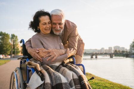 Senior woman in wheelchair walking with caregiver old man on road in park. Elderly family couple man supporting embracing woman in chair for people with disability outdoor. Rehabilitationの写真素材
