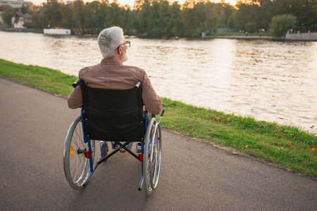 Sad senior man sitting in wheelchair walking on road in city park outdoor, back view. Old paralyzed man in chair for people with disability feel depressed. Lonely elderly sick man sits on wheelchairの写真素材