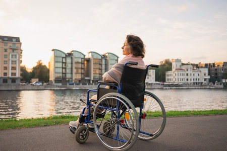 Sad senior woman sitting in wheelchair walking on road in city outdoor, back view. Old paralyzed lady in chair for people with disability feel depressed. Lonely elderly sick woman sits on wheelchairの写真素材