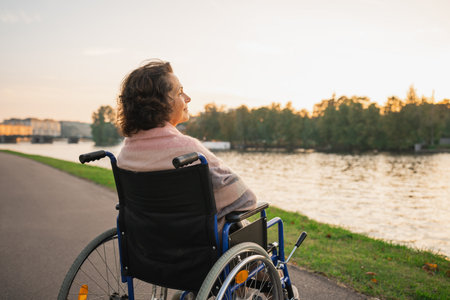 Sad senior woman sitting in wheelchair walking on road in city outdoor, back view. Old paralyzed lady in chair for people with disability feel depressed. Lonely elderly sick woman sits on wheelchairの写真素材