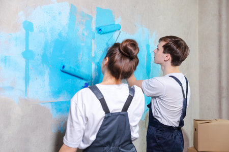 House decoration DIY renovation concept. Happy couple in empty room of new home painting wall together. Husband and wife holding paint roller painting wall with blue color paint in new houseの写真素材