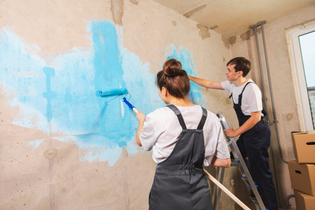 House decoration DIY renovation concept. Happy couple in empty room of new home painting wall together. Husband and wife holding paint roller painting wall with blue color paint in new houseの写真素材