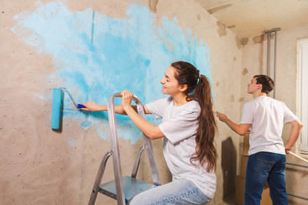 Couple in new home during repair works painting wall together. Happy family holding paint roller painting wall with blue color paint in new house. Home renovation DIY renew home conceptの写真素材