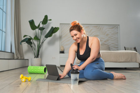 Yoga fitness workout training online. Woman practicing stretching with online app lessons at home. Plus size female doing sports exercise on yoga mat indoor. Girl have training workout for weight lossの写真素材