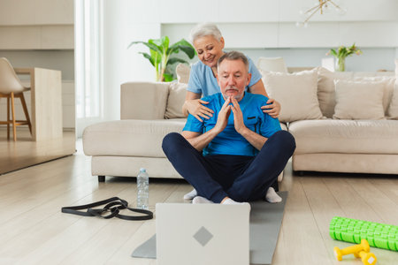 Yoga mindfulness meditation. Senior adult mature couple practicing yoga with online lessons in laptop. Old wife helping teaching husband meditating relaxing. Family doing breathing practice at homeの写真素材
