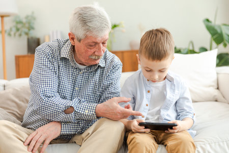 Happy family at home. Old senior man grandfather boy grandson enjoying time together watching video on phone. Good time at home. grandparent and child together. Older generation modern tech usageの写真素材