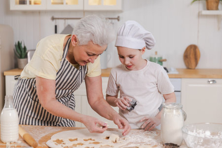 Happy family in kitchen. Grandmother granddaughter child cutting cookies of dough on kitchen table together. Grandma teaching kid girl cook bake cookies. Household teamwork helping family generationsの写真素材
