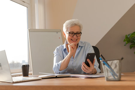 Confident stylish european middle aged senior woman using smartphone at workplace. Stylish older mature 60s gray haired lady businesswoman with cell phone in office. Boss leader using internet appsの写真素材