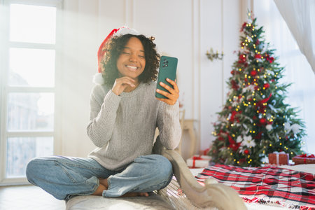 Merry Christmas. African woman in Santa hat having video chat in smartphone. Blogger in festive mood recording video in social network. Girl having video call with friends family celebrating Christmasの写真素材