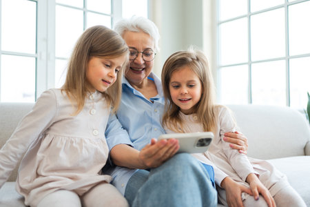 Happy family at home. Two little girls sisters twins grandmother enjoying time together watching video on phone. Good time at home. Grandma granddaughters child kids together. Family generationsの写真素材