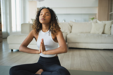 Yoga mindfulness meditation. Young healthy African girl practicing yoga at home. Woman sitting in lotus pose on yoga mat meditating relaxing indoor. Girl doing breathing practice. Yoga at homeの写真素材